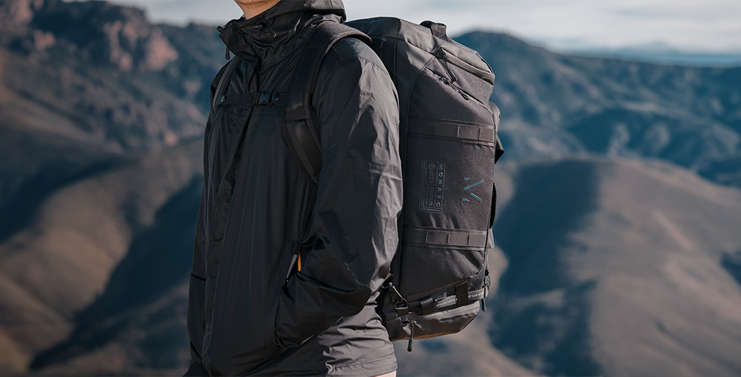 A hiker outfitted in a dark jacket carries a Monarc backpack while standing against a stunning mountainous backdrop. The backpack, prominently featuring the Monarc logo.