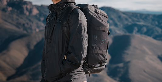 A hiker outfitted in a dark jacket carries a Monarc backpack while standing against a stunning mountainous backdrop. The backpack, prominently featuring the Monarc logo.