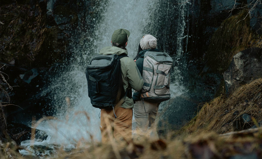 Two hikers wearing Monarc duffel backpacks stand in front of a cascading waterfall, surrounded by lush greenery and rugged rocks, showcasing the gear's durability and suitability for outdoor adventures.