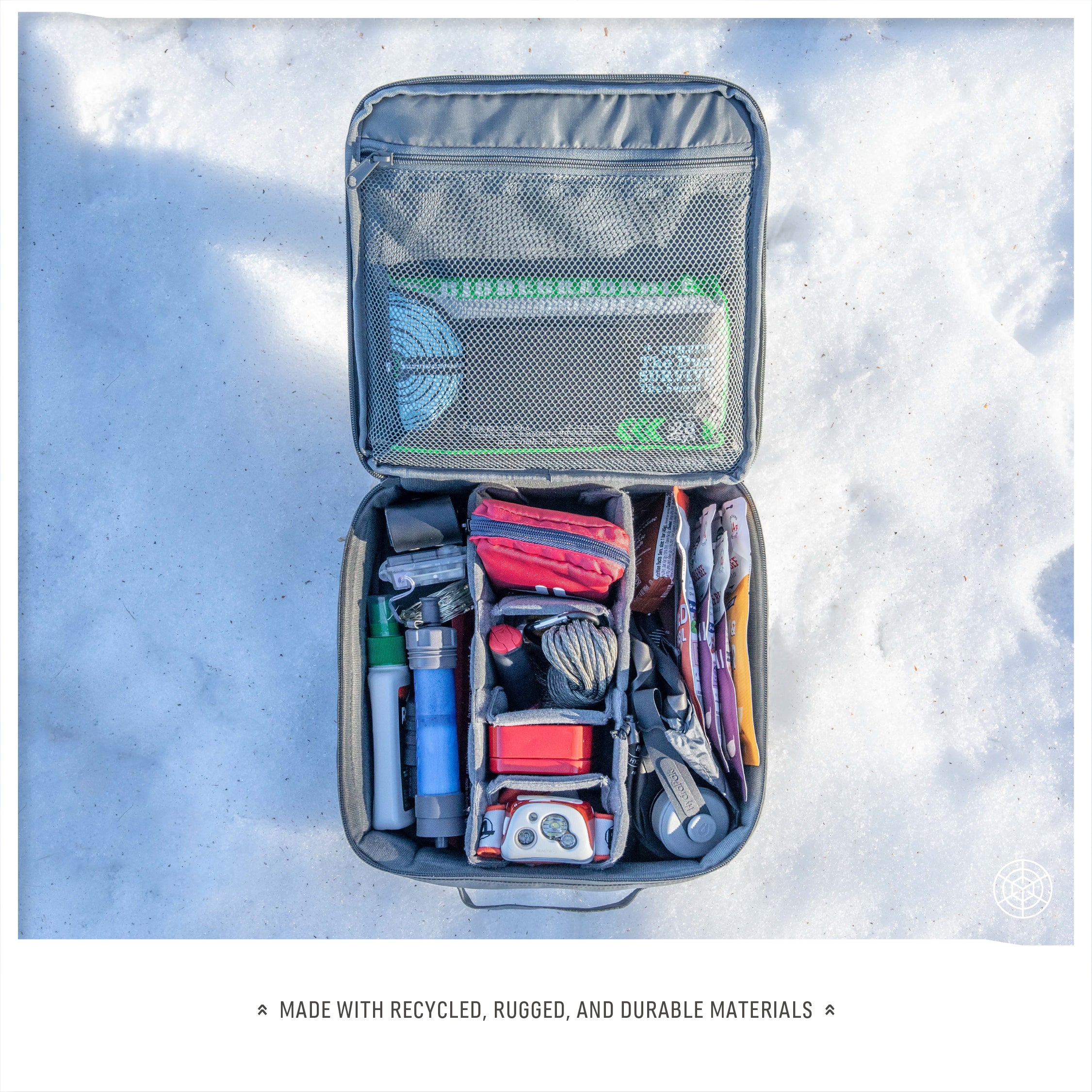  Outdoor photo showing the Monarc Camera Cube in gray, open on snowy ground, packed with a diverse array of items including tools, stationery, and survival gear. The cube's versatility is highlighted by its capacity to organize a variety of essentials beyond photography, crafted from durable, recycled materials suited for rugged use.